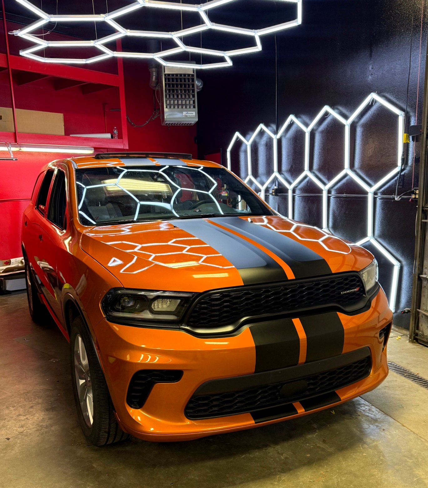 Orange Dodge Durango with matte black vinyl stripes under hexagon lights